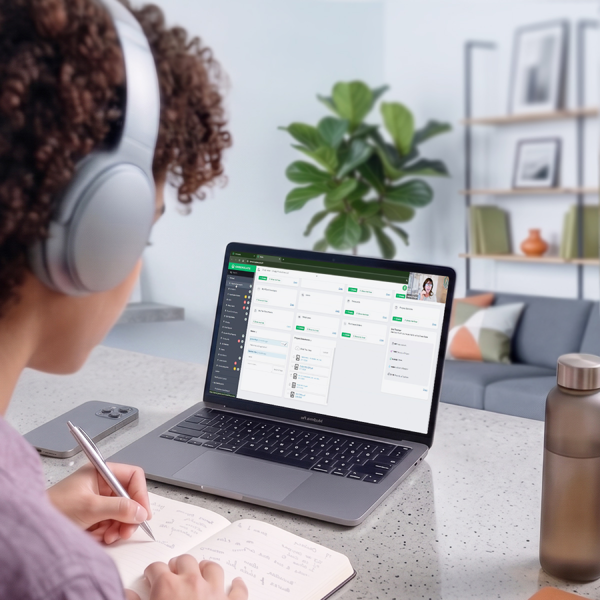 "A person wearing headphones writes in a notebook while a laptop shows a project dashboard with a video call, in a softly lit home setting with a plant and sofa in the background."